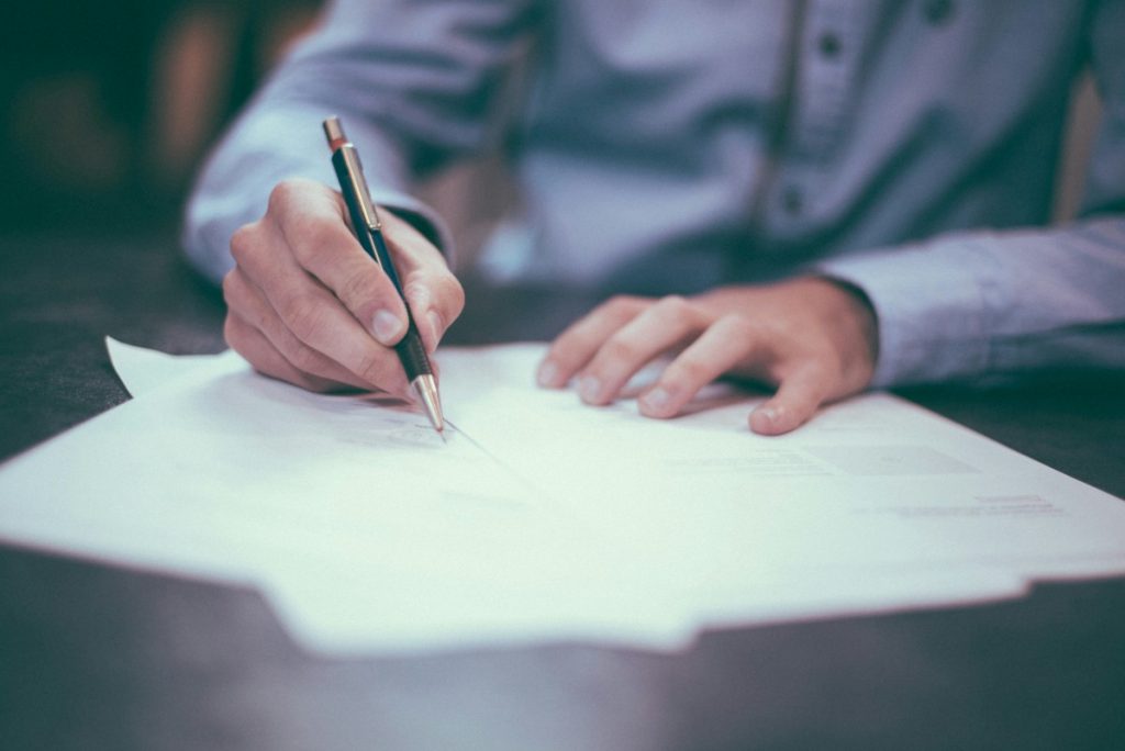 Cette image représente une personne en train de rédiger ou signer un document officiel. Le focus est mis sur la main tenant un stylo et les feuilles de papier sur une table, ce qui suggère un contexte administratif ou contractuel, comme la signature d'un contrat de domiciliation. L'arrière-plan est flou, dirigeant toute l'attention vers l'action d'écriture.