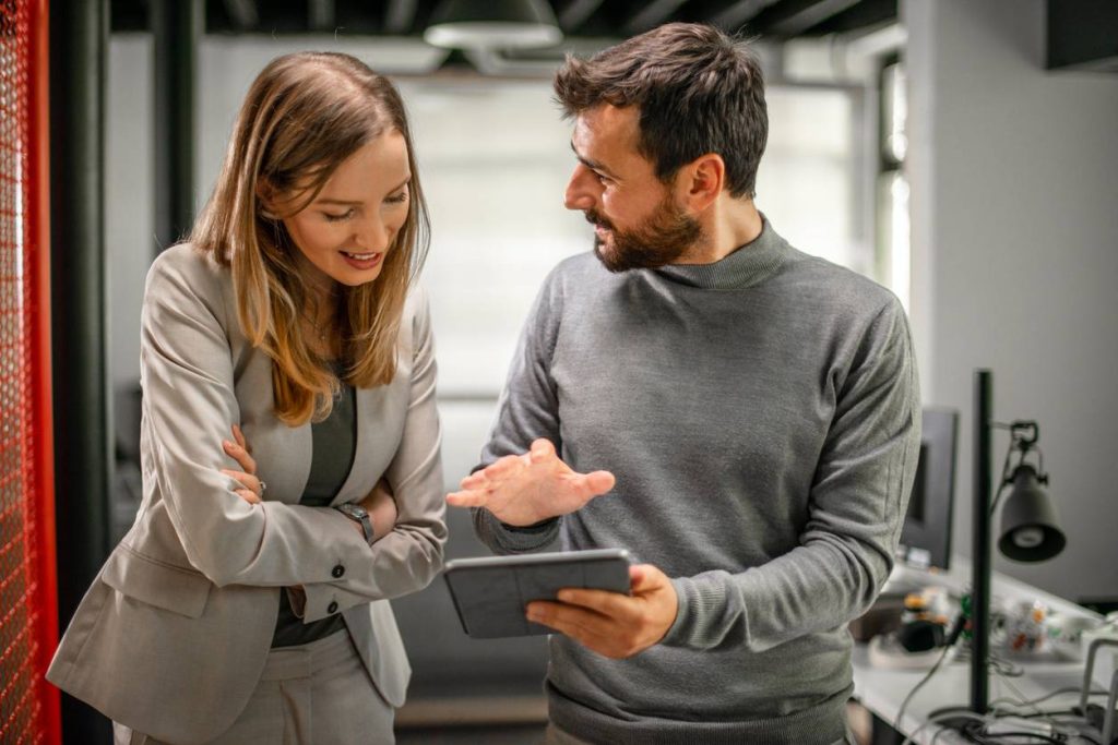 Un homme et une femme en discussion dans un bureau moderne, regardant une tablette numérique. L'homme explique quelque chose avec un geste de la main tandis que la femme écoute avec intérêt.